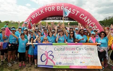 A large group of event participants stand under the finish line holding a sign for Capital Breast Care Center