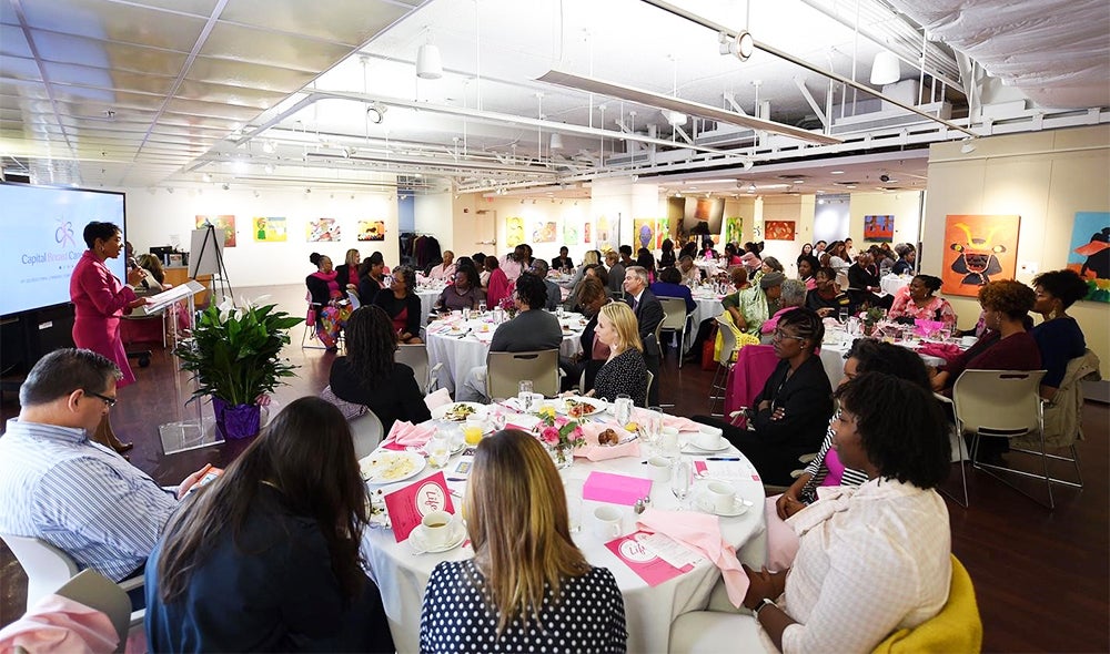 A group of people are seated at tables in a large room; a speaker stands at a podium