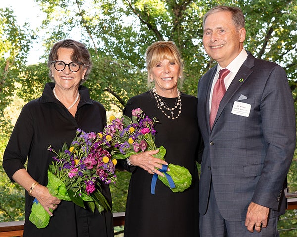 The co-chairs stand together holding bouquets of flowers next to Dr. Weiner