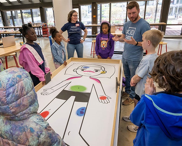 Becca and a KID Museum staffer speak with a group of children about the drawing of a person that is spread on the table before them