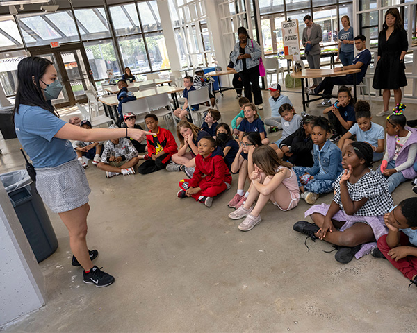 A KID Museum staffer addresses a large group of students seated on the floor in front of her