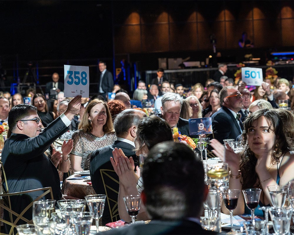 People are seated at tables eating and drinking while some hold up signs to participate in the live appeal