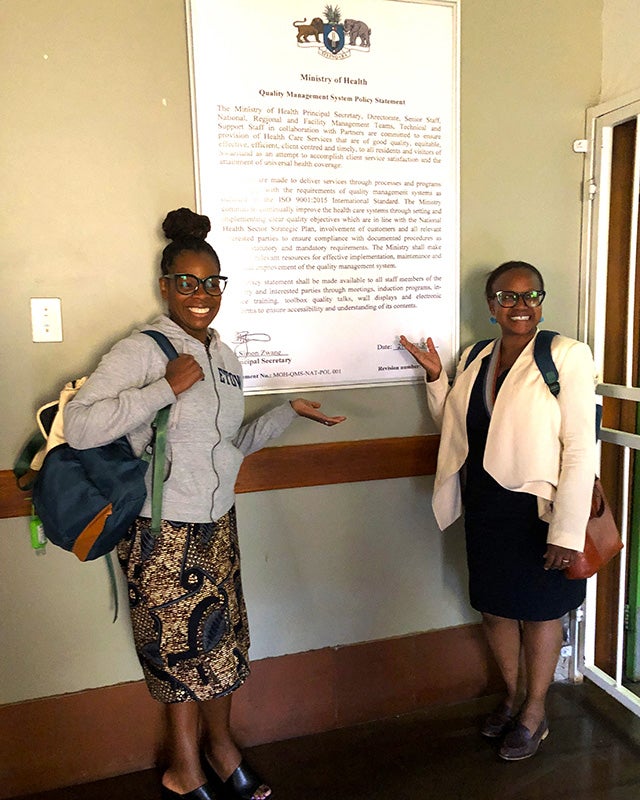 Two women stand before a sign detailing the Ministry of Health Quality Management System Policy Statement
