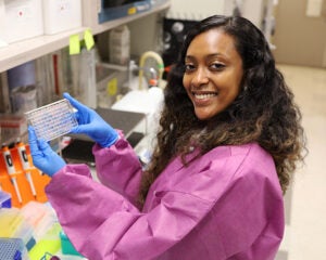 A young Black woman in pink scrubs and blue plastic gloves holds a piece of lab equipment and smiles at the camera