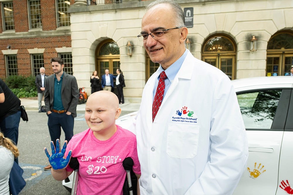 A young cancer patient with crutches holds up her painted hand while standing with Dr. Uren near a Hyundai vehicle