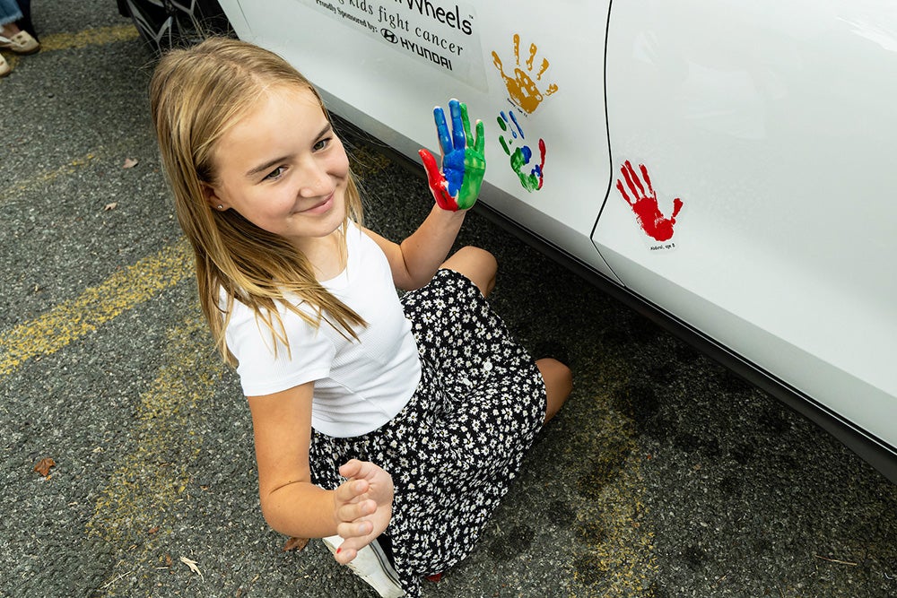 Emmy holds up her painted palm after making a painted handprint on the side of a white Hyundai vehicle