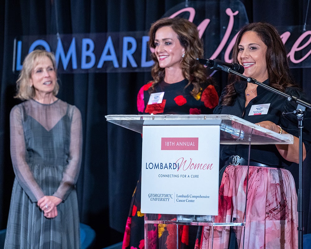 Three women stand  behind a podium onstage at Lombardi Women