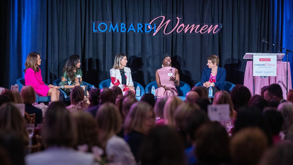 Panelists sit and talk onstage in chairs while the attendees at Lombardi Women look on