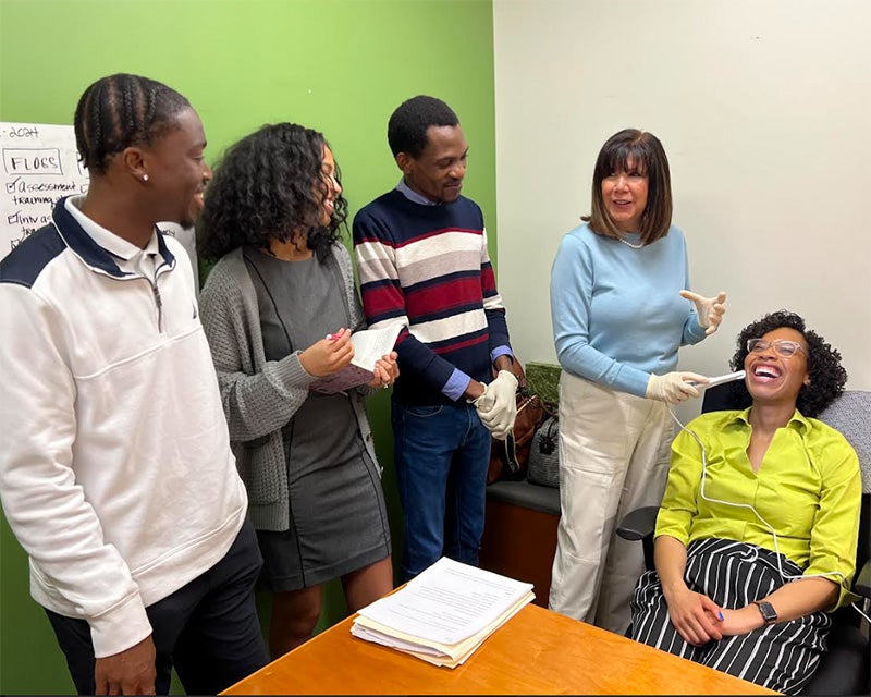 Four people stand, one holding a device to the mouth of a Black woman seated in a chair