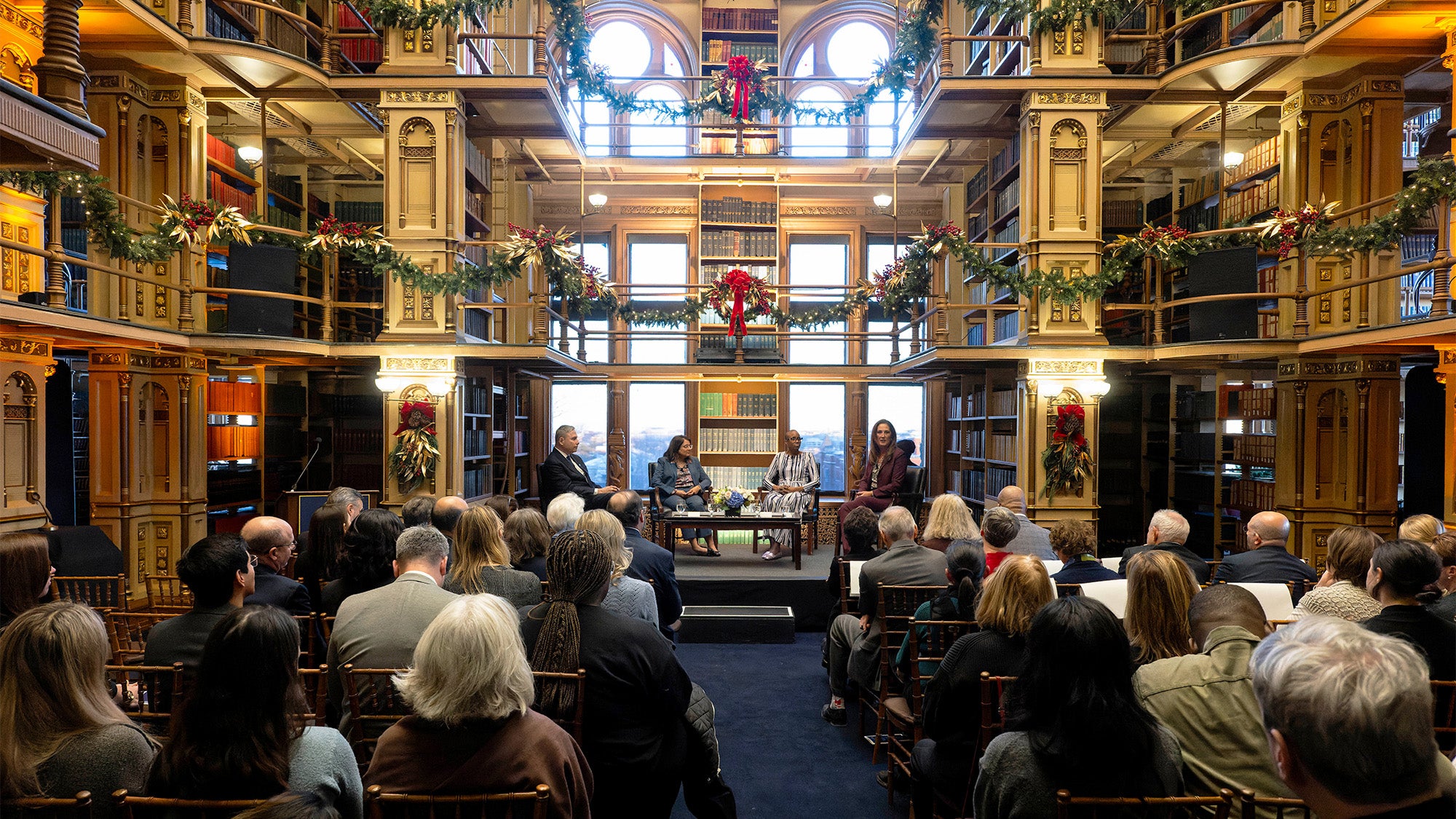 A long view of the speakers on a stage in Riggs Library seated before an audience in rows of chairs