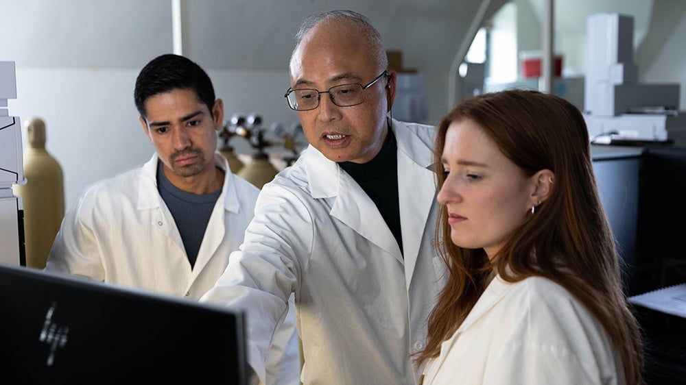 Two students and a researcher in white coats stand together looking at a computer screen