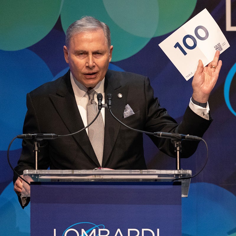 Lou Weiner stands behind a podium and holds up a sign that reads 100