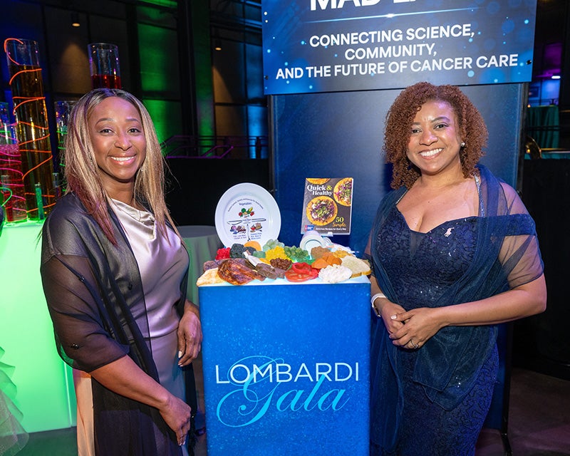 Mireille and Natalie from Ralph Lauren Center stand with plastic models of food and a plate printed with portion sizes of various food groups