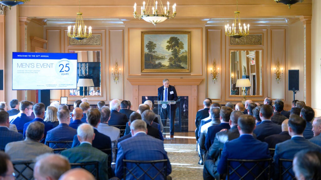 Dr. Weiner speaks to a large room full of people from a podium with a screen next to him displaying the words Men's Event 2025