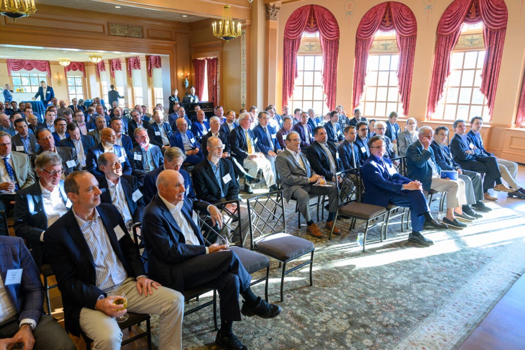 A roomful of men seated in rows of chairs in a large ballroom listens to a speaker