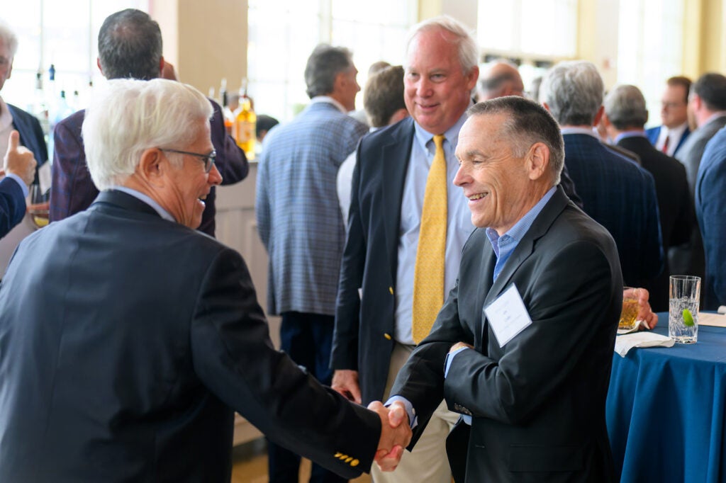 Two men shake hands as another looks on smiling amid a room of people
