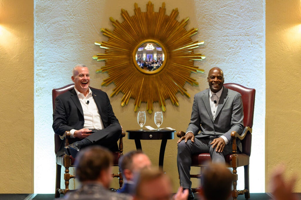 Two men sit in chairs onstage before an audience