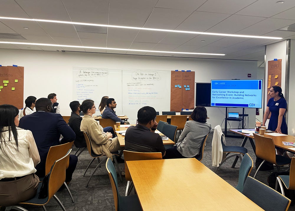 People sit at tables as a presenter stands at the front of the room next to a monitor