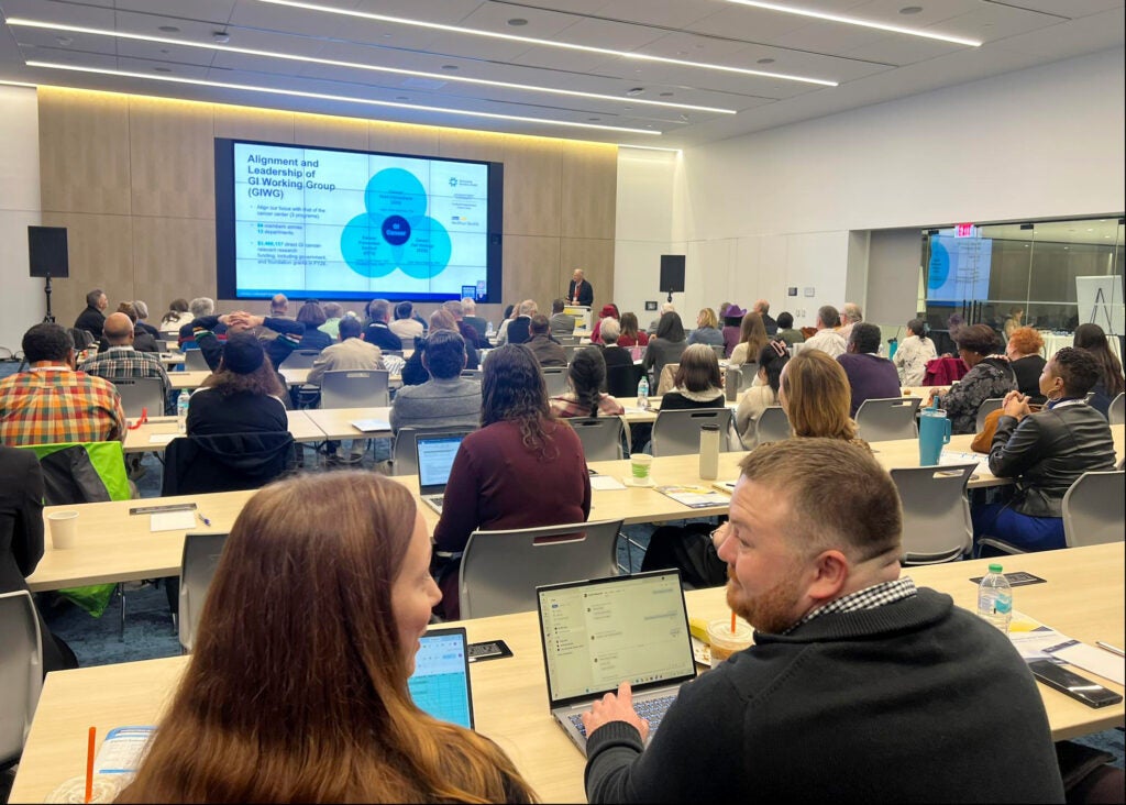 A conference room full of attendees listens to a presentation at Day 2 of the Ruesch Symposium