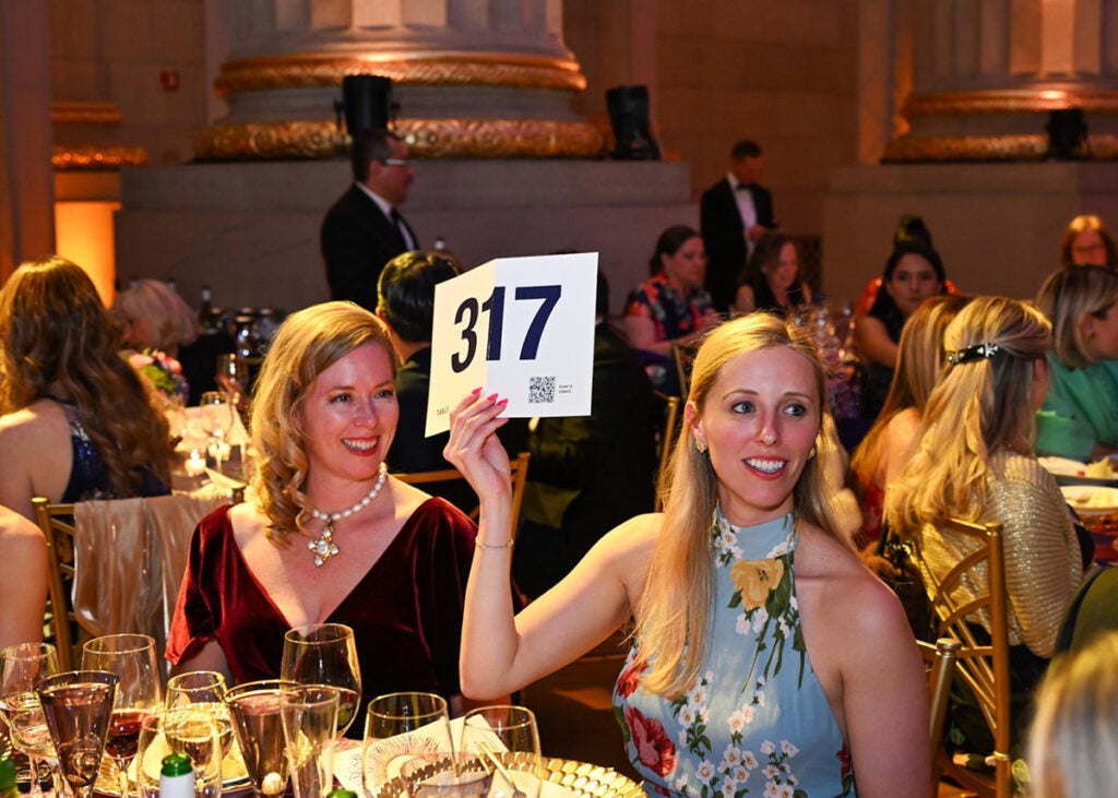 Two women sit at a table, one holding up a sign to bid in the live appeal