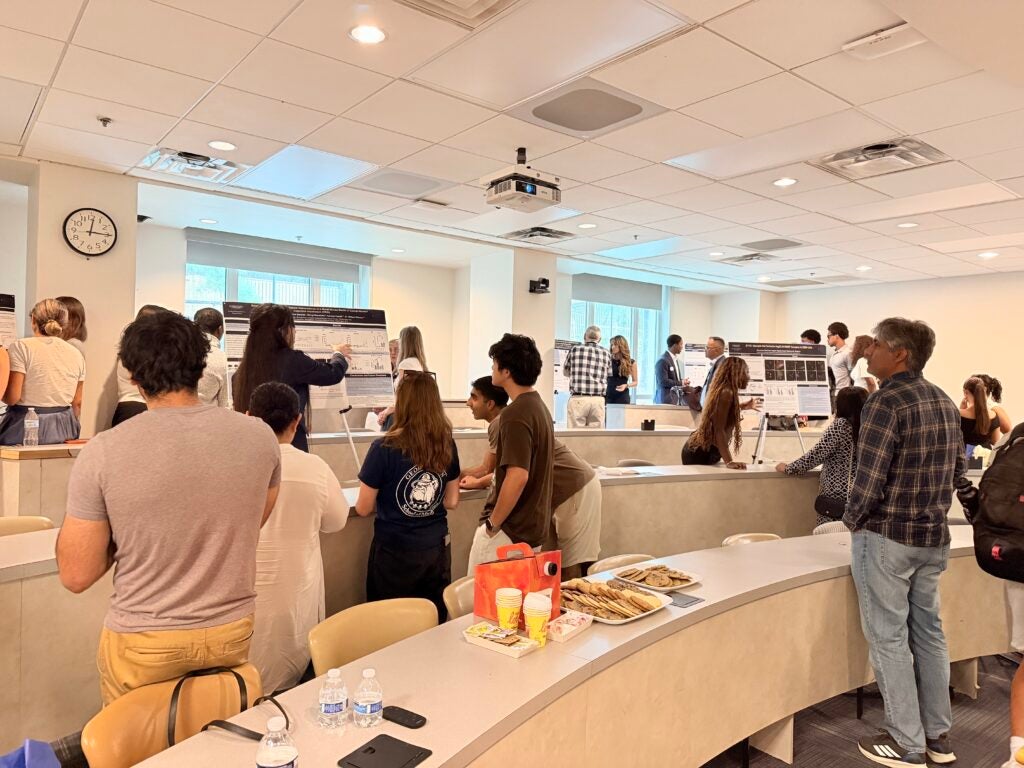 Visitors walking around in a classroom with during a CURE-M poster presentation and students standing by their posters.