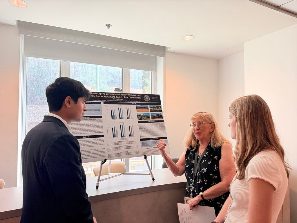 A young man by a his research poster and two women appear to ask questions about his poster